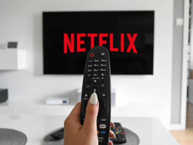 Mano sosteniendo control remoto apuntando a logo de Netflix en pantalla. Servicios de plataformas digitales de streaming afectados por la Reforma Fiscal Plataformas Digitales en México.