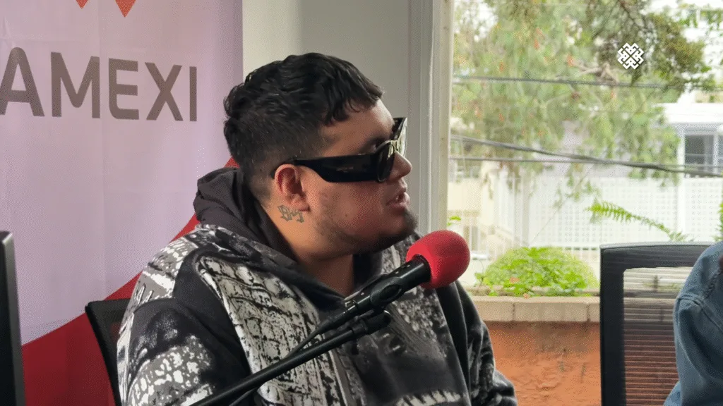 Primer plano de Loojan, integrante de Cachirula & Loojan, usando gafas de sol y hablando en un micrófono rojo durante la entrevista sobre 'Sexolandia'.