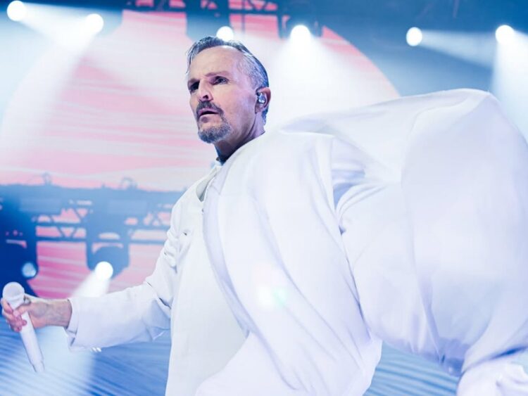 Miguel Bosé cantando en el escenario durante un concierto, iluminado por luces de espectáculo.