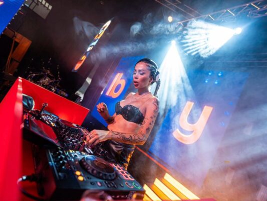 DJ mexicana tocando en un escenario masivo de música electrónica ante una multitud durante un festival nocturno.
