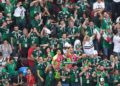 Aficionados mexicanos celebran durante un partido en una zona de reunión deportiva con banderas y camisetas de la selección.
