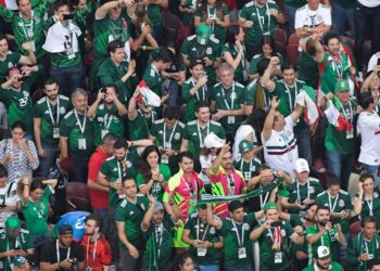 Aficionados mexicanos celebran durante un partido en una zona de reunión deportiva con banderas y camisetas de la selección.