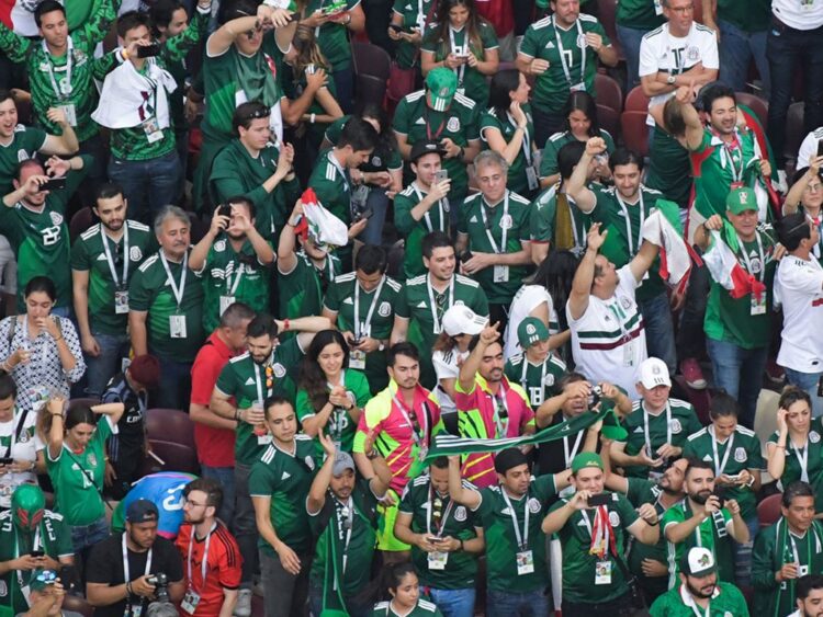 Aficionados mexicanos celebran durante un partido en una zona de reunión deportiva con banderas y camisetas de la selección.