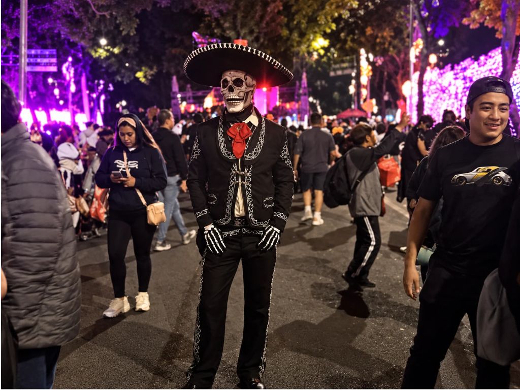 Persona vestida de charro negro con sombrero y maquillaje de calavera durante una celebración nocturna de Día de Muertos, entre luces de colores y asistentes disfrazados.