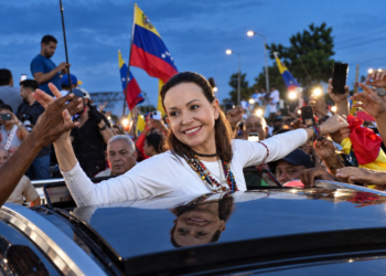 Mujer de blanco saluda a una multitud desde un vehículo, rodeada de simpatizantes con banderas de Venezuela.